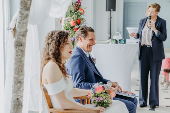 Freie Rednerin spricht bei einer Zeremonie drinnen, das Brautpaar sitzt nebeneinander mit Blumenstrauß.