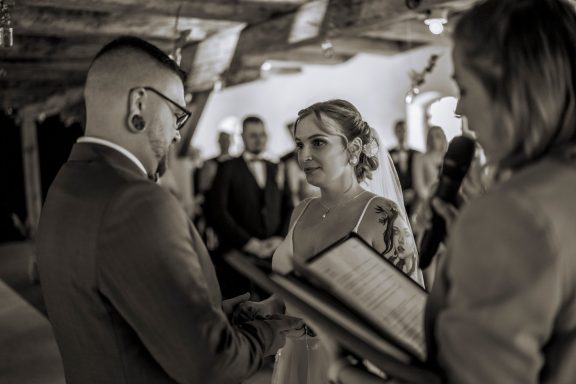 Freie Rednerin spricht zu einem emotional bewegten Brautpaar während einer Trauung im rustikalen Innenraum.