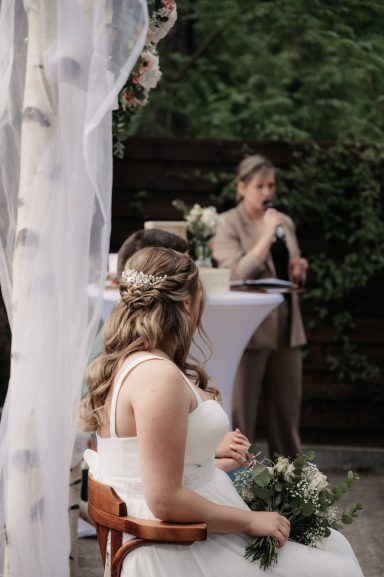 Braut sitzt mit Blumen im Haar und hört der Rednerin bei der Zeremonie zu.