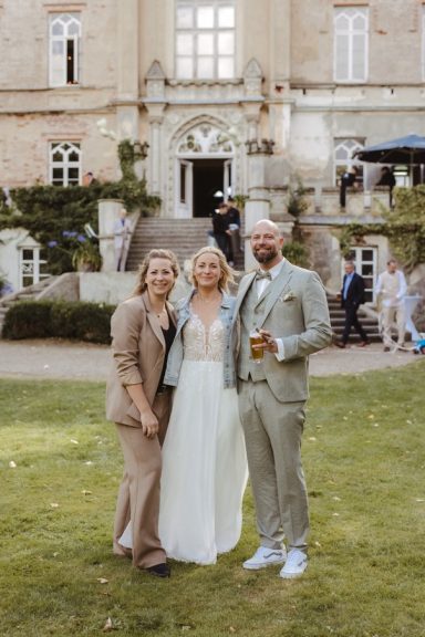 Rednerin und Brautpaar stehen zusammen vor einem alten Herrenhaus und lächeln in die Kamera