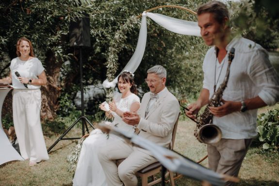 Brautpaar sitzt vor Traubogen und feiert die Musik eines Saxophonisten